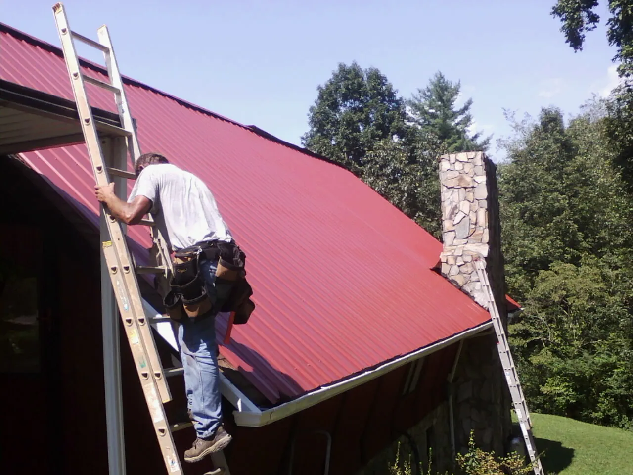 Roofer on a ladder beside a red metal roof and stone chimney — Soffit & Fascia Repair by Roofing Brothers in La Verne